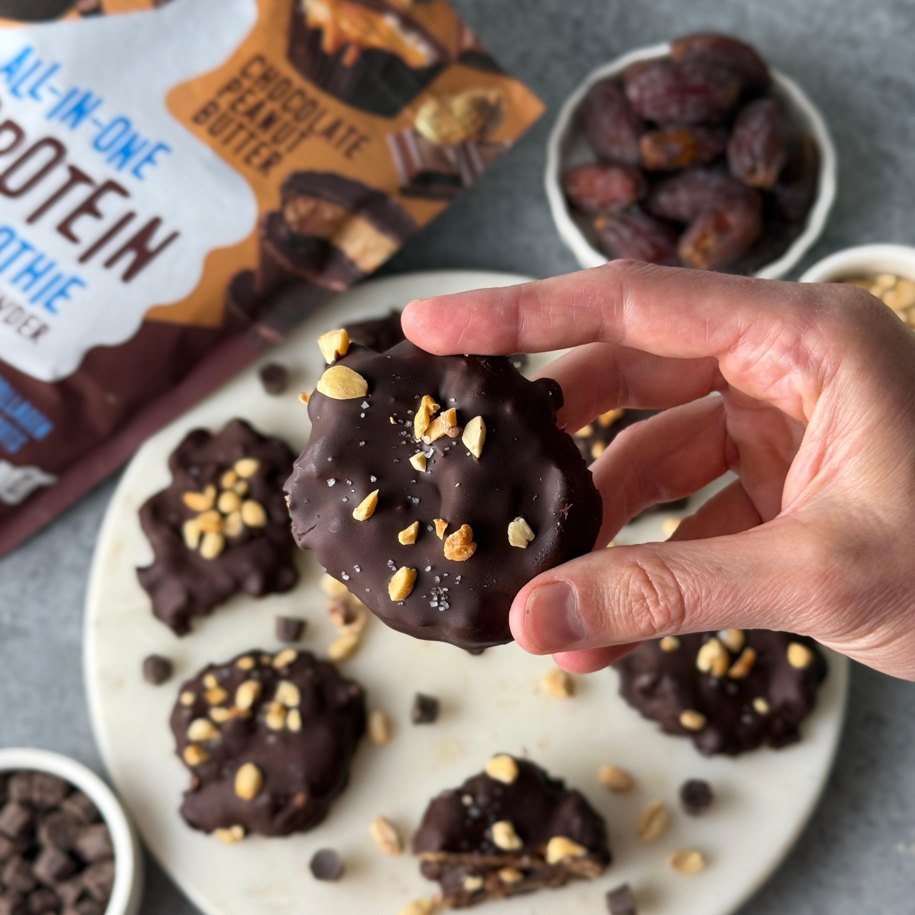 Hand holding a chocolate-covered protein cookie topped with chopped peanuts and sea salt, surrounded by similar cookies, dates, chocolate chips, and FlavCity protein smoothie powder packaging.