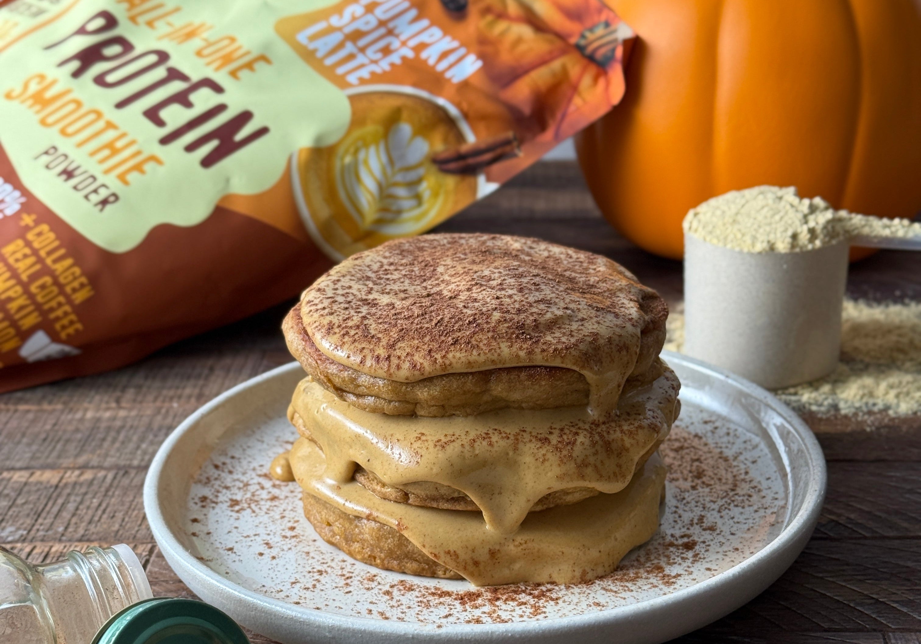 Stack of fluffy pumpkin spice pancakes topped with creamy sauce, surrounded by FlavCity protein powder pouch, pumpkin, measuring scoop, and jar of pumpkin spice.