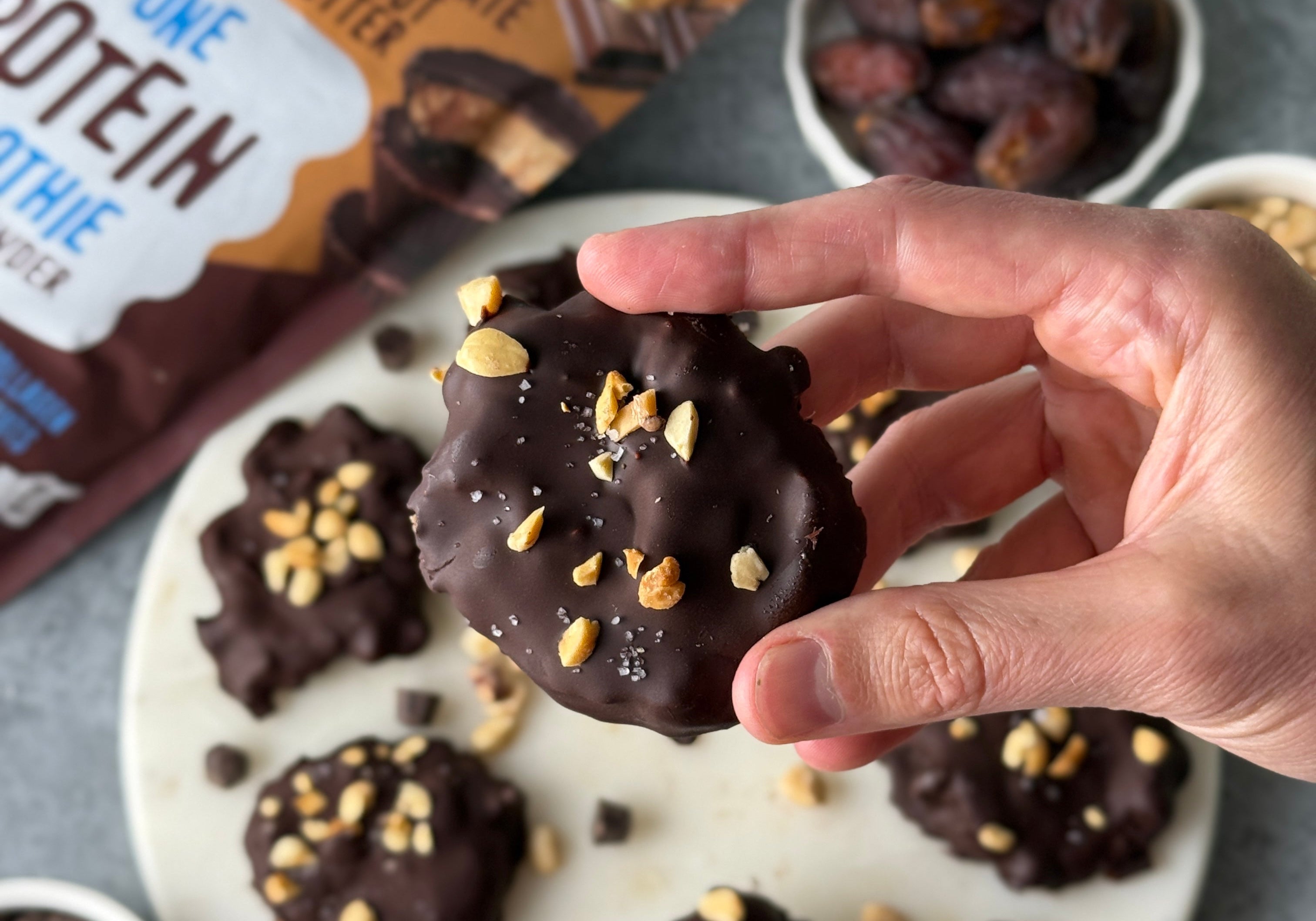 Hand holding a chocolate-covered protein cookie topped with chopped peanuts and sea salt, surrounded by similar cookies, dates, chocolate chips, and FlavCity protein smoothie powder packaging.
