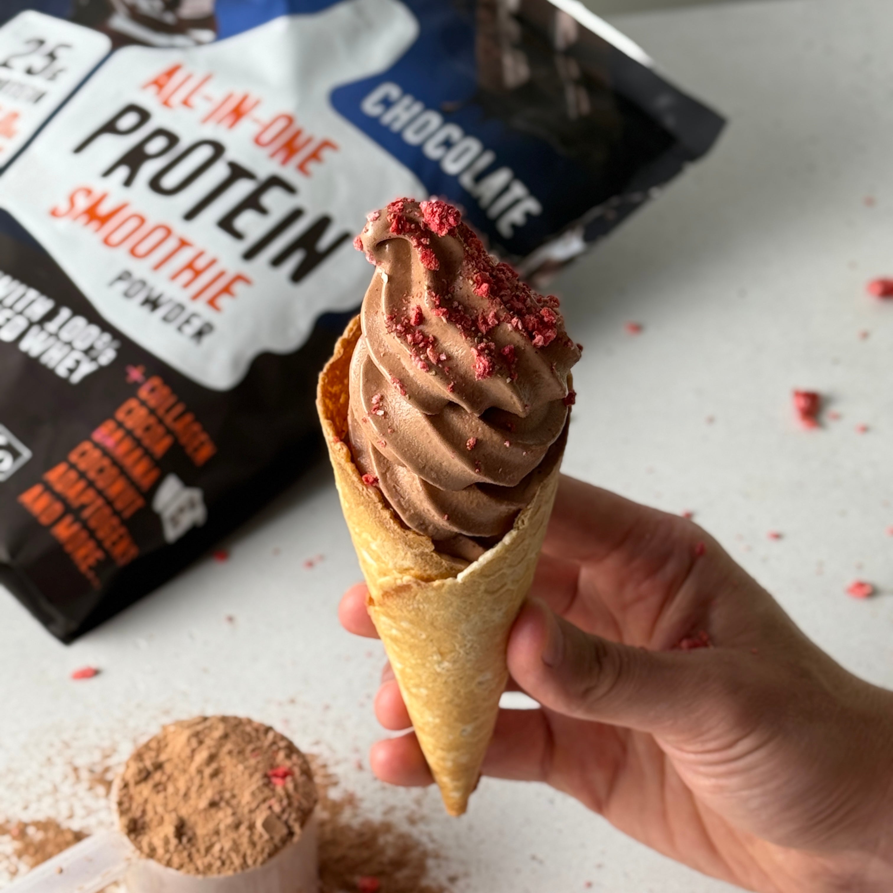 Hand holding a chocolate protein smoothie ice cream cone, topped with crushed berries, with FlavCity Chocolate Protein Smoothie Powder and scoop in the background on a white counter.