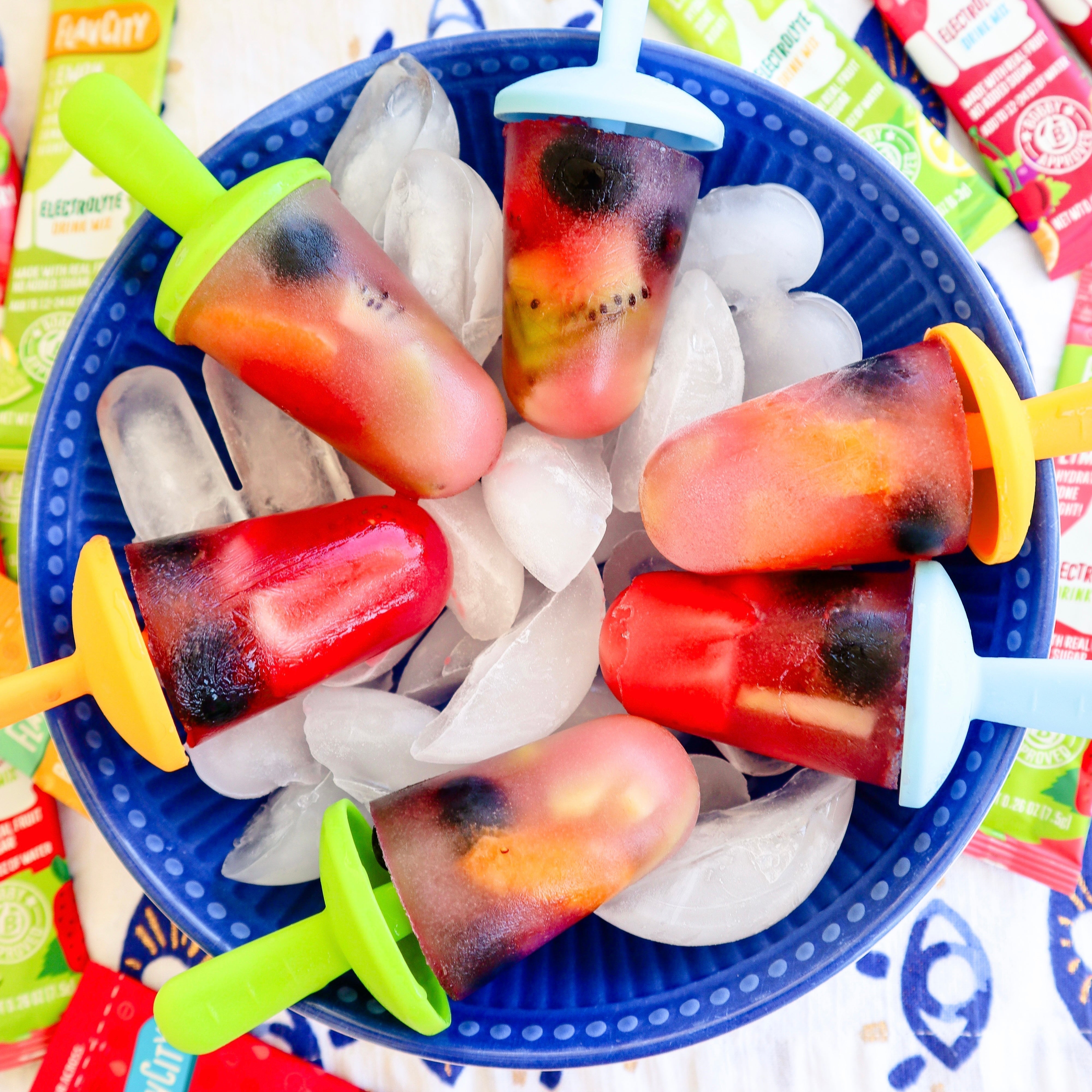 Colorful homemade fruit popsicles with visible slices of kiwi, blueberries, and other fruits, served in a blue bowl filled with ice, surrounded by FlavCity Electrolyte mix packets