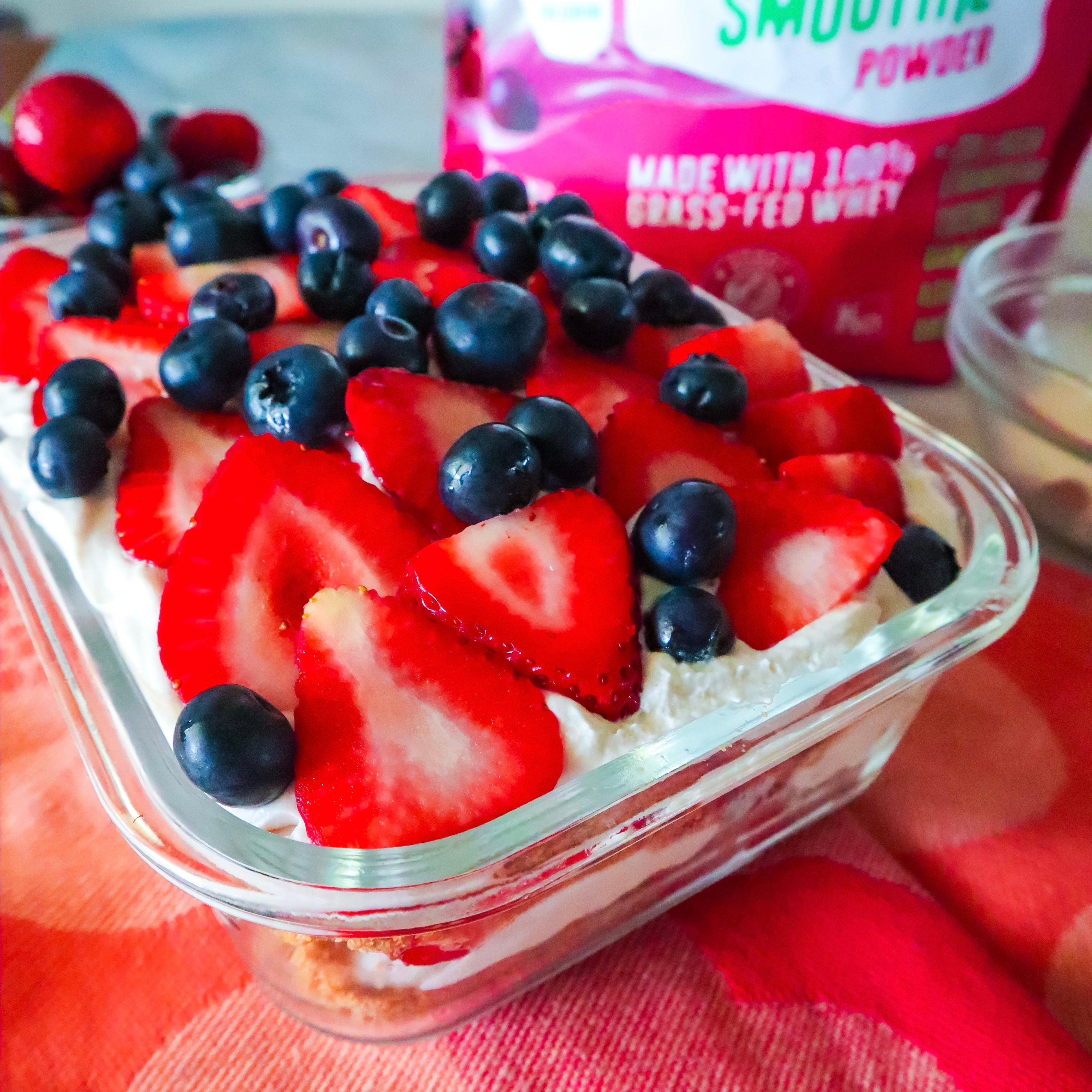 Berry cake dessert topped with fresh sliced strawberries and blueberries in a glass dish, sitting on a red cloth, with smoothie whey powder in the background.