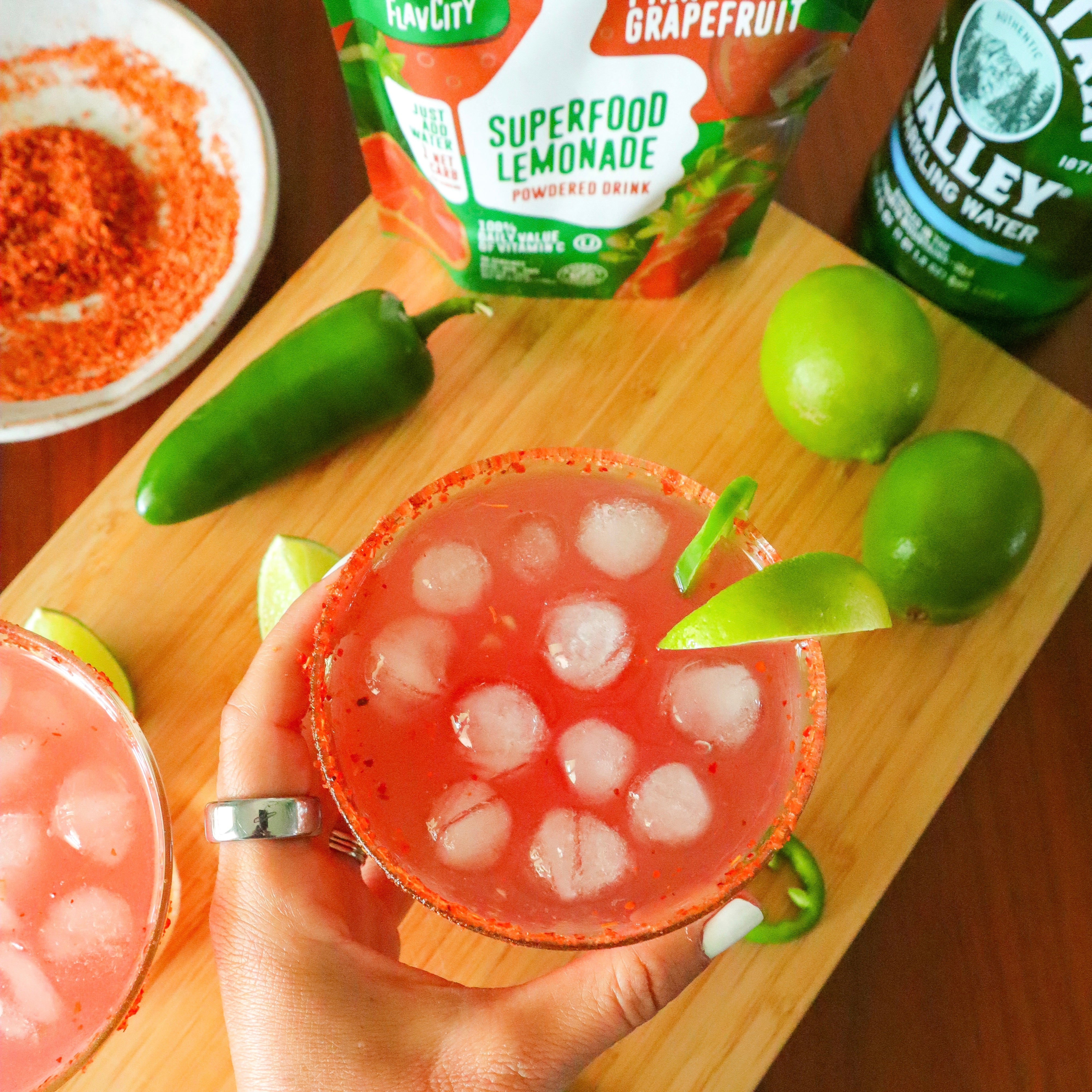 Refreshing grapefruit lemonade cocktail with ice, garnished with a lime wedge and sliced jalapeño, on a wooden board next to limes, jalapeño, and superfood drink mix.