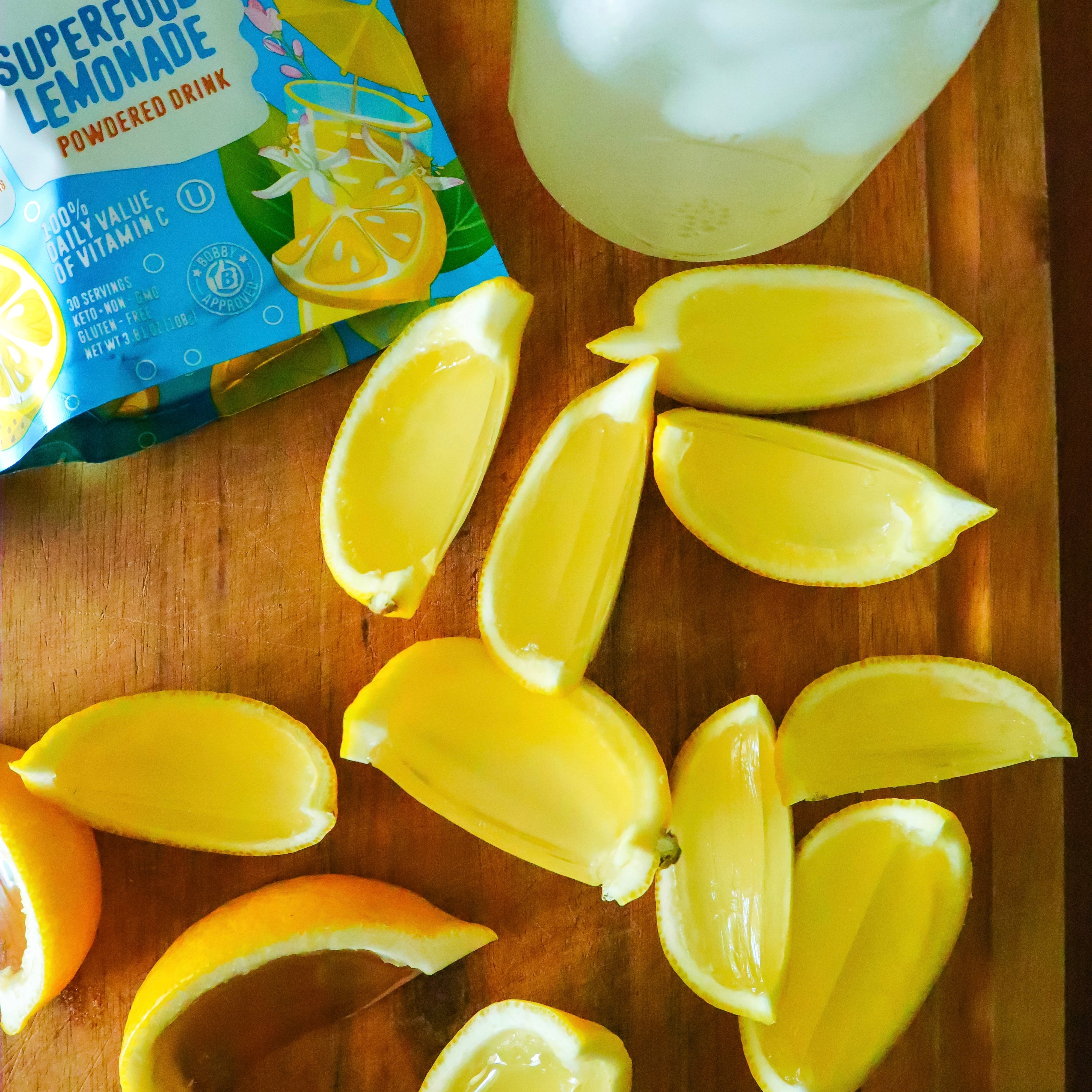 Sliced lemon wedges with pulp removed are arranged on a wooden board next to a packet of superfood lemonade powder and a mason jar of iced lemonade.