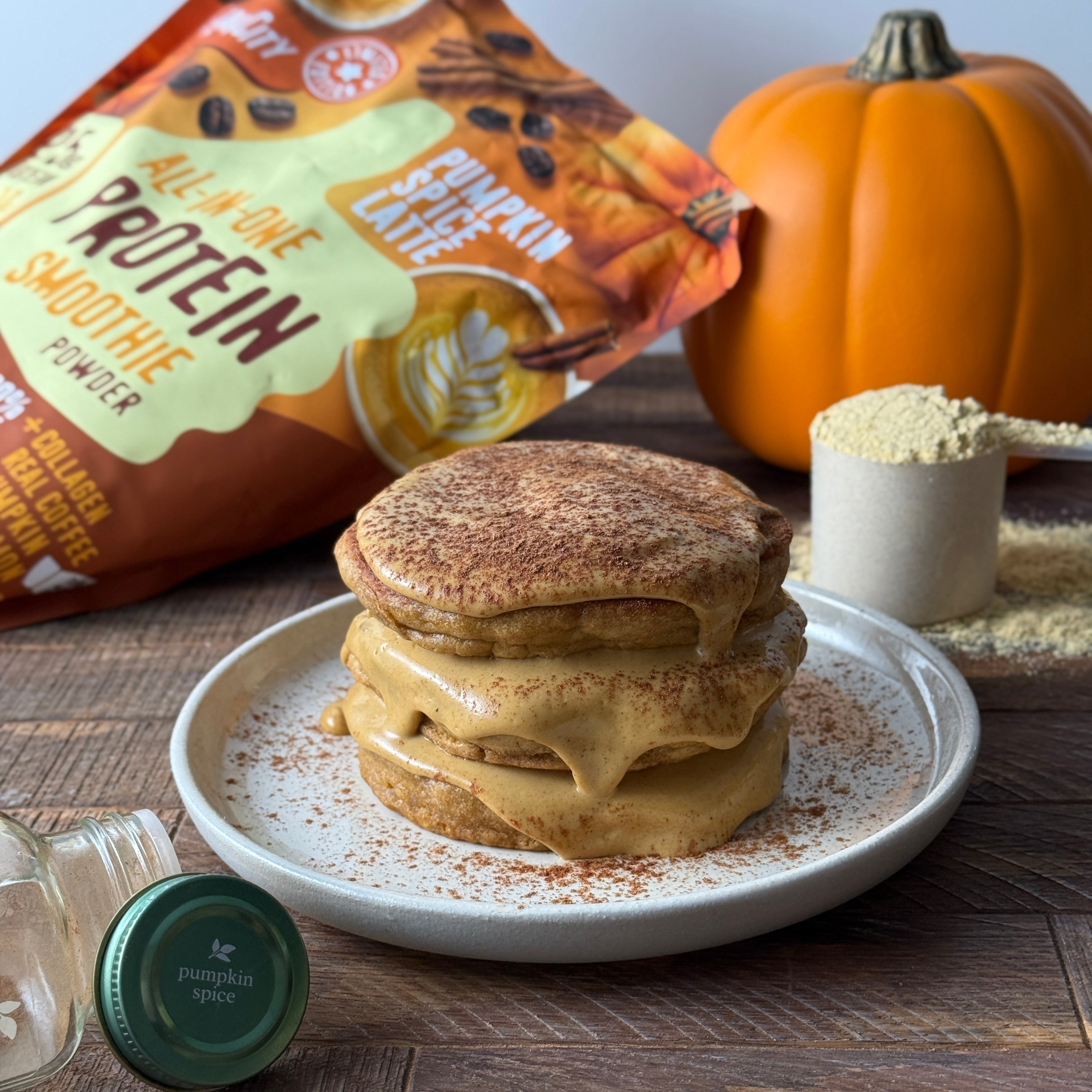 Stack of fluffy pumpkin spice pancakes topped with creamy sauce, surrounded by FlavCity protein powder pouch, pumpkin, measuring scoop, and jar of pumpkin spice.