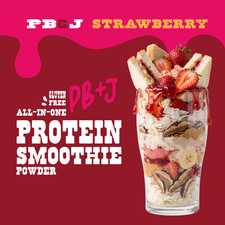 A Shop FlavCity PB&J Strawberry Protein Smoothie in a tall glass layered with protein powder, peanut butter, sliced strawberries, banana, and coconut flakes, topped with strawberry jam and a miniature PB&J smoothie sandwich.