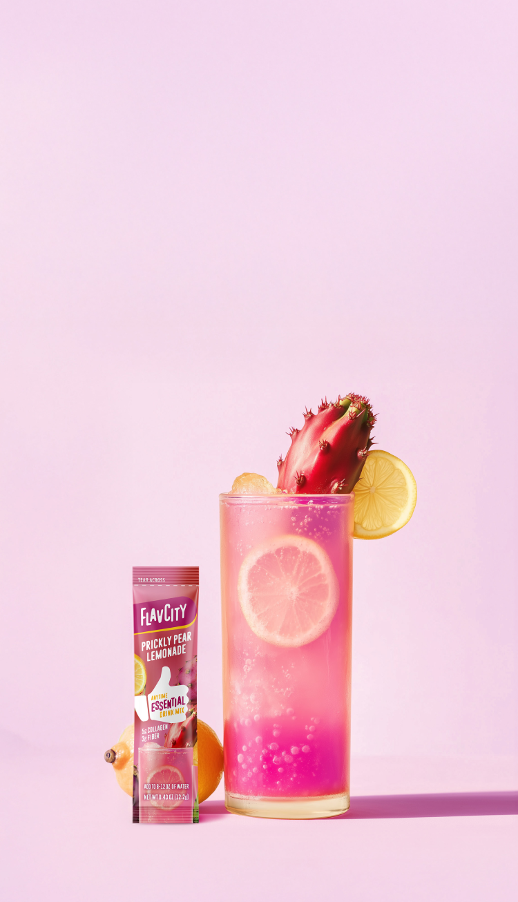 A glass of pink prickly pear lemonade with lemon slices and a prickly pear garnish is beside a FlavCity Prickly Pear Lemonade drink mix packet, all set against a pastel pink background.