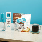 A bag of Shop FlavCity Coffee With Benefits Medium Roast, a blue handheld milk frother with its box, and a glass cup of black coffee all sit on a countertop against a blue background.