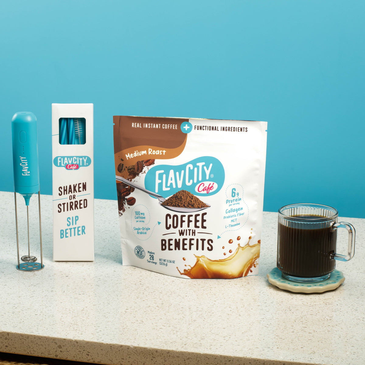 A bag of Shop FlavCity Coffee With Benefits Medium Roast, a blue handheld milk frother with its box, and a glass cup of black coffee all sit on a countertop against a blue background.
