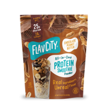A stand-up pouch of Shop FlavCity Chocolate Peanut Butter Protein Smoothie powder next to a glass filled with a smoothie topped with banana slices, nuts, and chocolate shavings.
