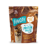 A stand-up pouch of Shop FlavCity Chocolate Peanut Butter Protein Smoothie powder next to a glass filled with a smoothie topped with banana slices, nuts, and chocolate shavings.