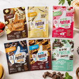 Six Shop FlavCity All-In-One Protein Smoothie Packets are arranged on a table with chocolate pieces and strawberries, showcasing flavors like Butter Coffee, Vanilla Cream, and Chocolate, and featuring grass-fed whey protein.