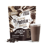 A bag of Shop FlavCity Cookies & Cream Protein Smoothie stands behind a glass of chocolate smoothie and some cookies scattered on a white surface.