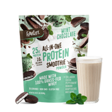 A pouch of Shop FlavCity Mint Chocolate Protein Smoothie stands behind a filled glass, with mint leaves and chocolate pieces nearby on a white background.