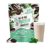 A pouch of Shop FlavCity Mint Chocolate Protein Smoothie stands behind a filled glass, with mint leaves and chocolate pieces nearby on a white background.