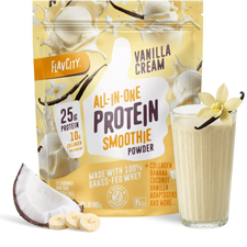 A pouch of Shop FlavCity Vanilla Cream Protein Smoothie sits next to a glass of vanilla protein smoothie, banana slices, coconut, and a vanilla flower, highlighting its grass-fed whey content and premium ingredients.
