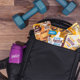 A Shop FlavCity Protein Smoothies Starter Kit, featuring a black gym bag filled with single-serve meal packets and a stainless steel shaker bottle in the pocket, sits next to two blue dumbbells on a wooden floor.
