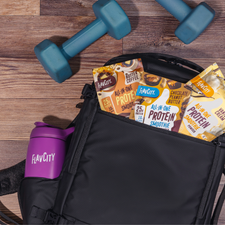 A Shop FlavCity Protein Smoothies Starter Kit, featuring a black gym bag filled with single-serve meal packets and a stainless steel shaker bottle in the pocket, sits next to two blue dumbbells on a wooden floor.