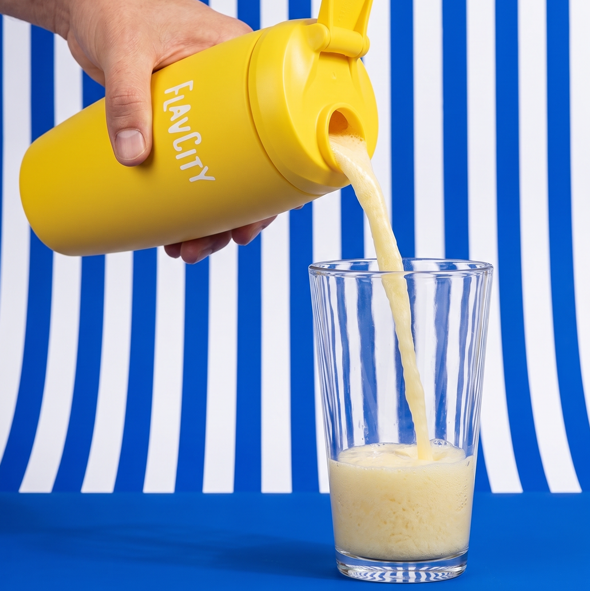 A hand pours a creamy yellow Shop FlavCity Whipped Lemonade Protein Smoothie from a branded bottle into a clear glass, set against a blue and white striped backdrop with a blue surface.