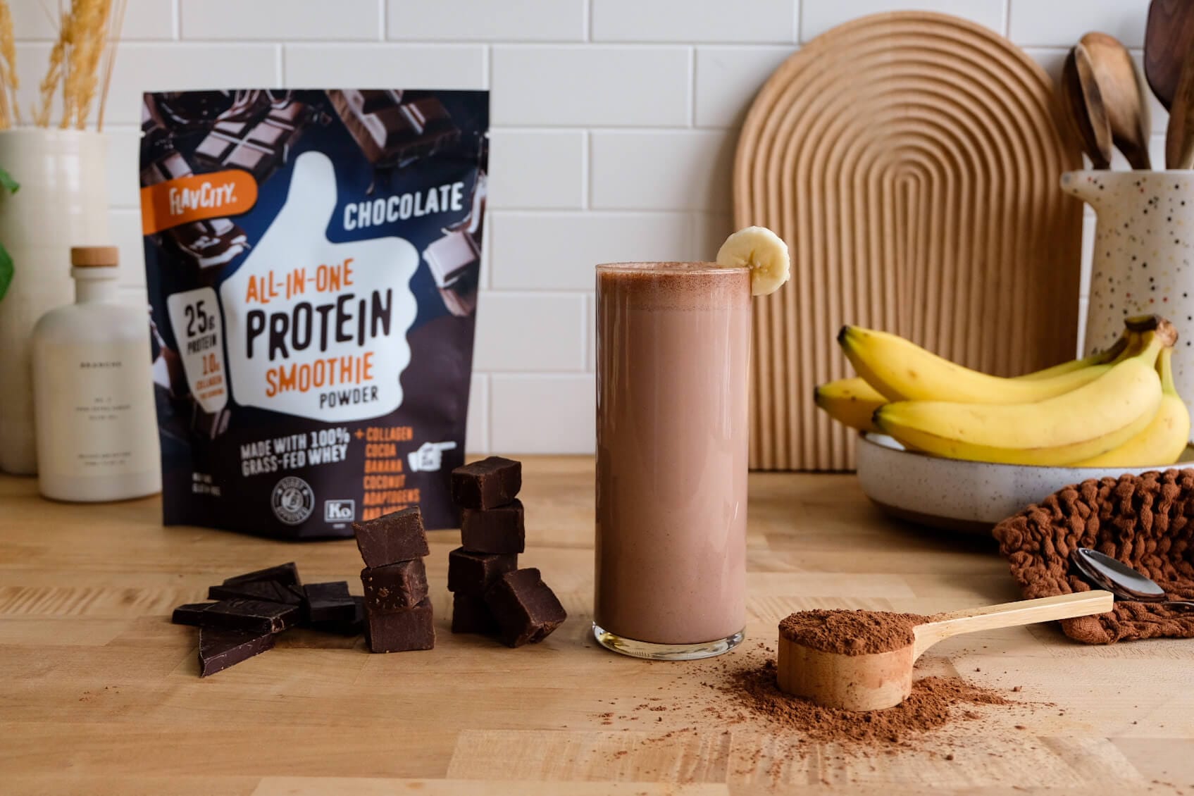 A glass of chocolate protein smoothie garnished with a banana slice sits on a wooden counter, surrounded by chocolate chunks, a scoop of cocoa powder, bananas, and a bag of chocolate protein powder.