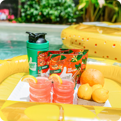 FlavCity Everyday Essentials in cups with a green bottle and orange slices on a pool float.