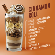 The Shop FlavCity Cinnamon Roll Protein Smoothie is served in a tall glass and topped with whipped cream, a cinnamon roll, banana, oats, and cinnamon. Ingredients include whey protein, collagen, cinnamon, banana, and coconut milk.